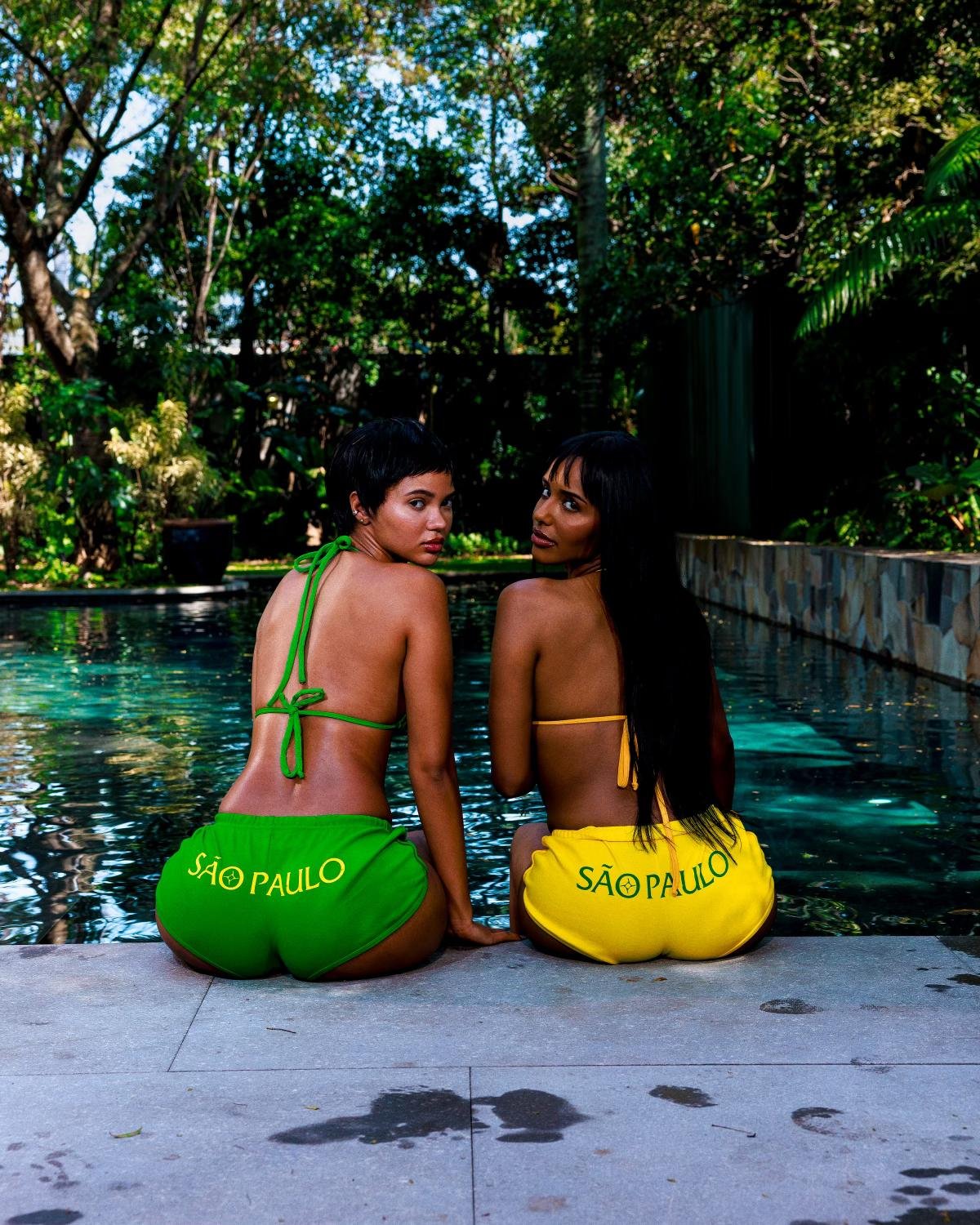 Duas mulheres sentadas à beira da piscina usam biquínis e shorts curtos nas cores verde e amarelo, estampados com \