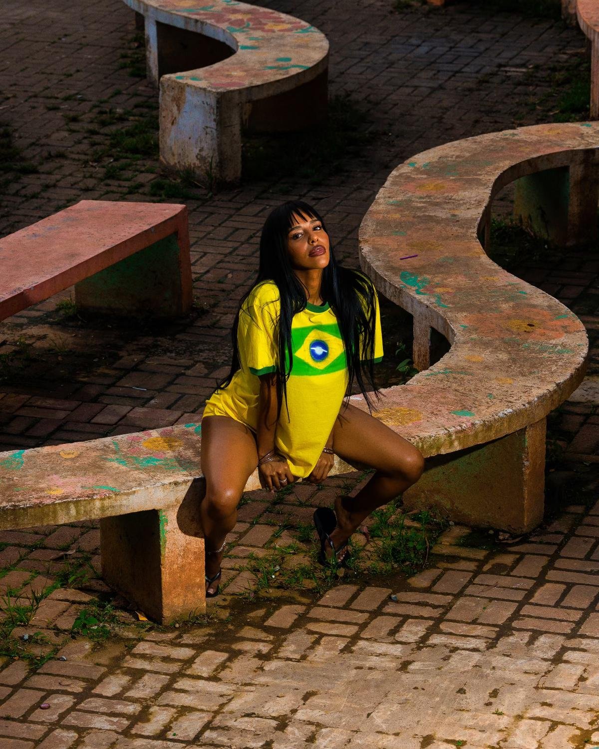 Mulher com camiseta oversized da seleção brasileira, uma tendência de moda casual e descolada. A camiseta amarela, com o emblema do Brasil, é usada de forma descontraída, unindo estilo esportivo e urbano. Foto em área externa com bancos de concreto.