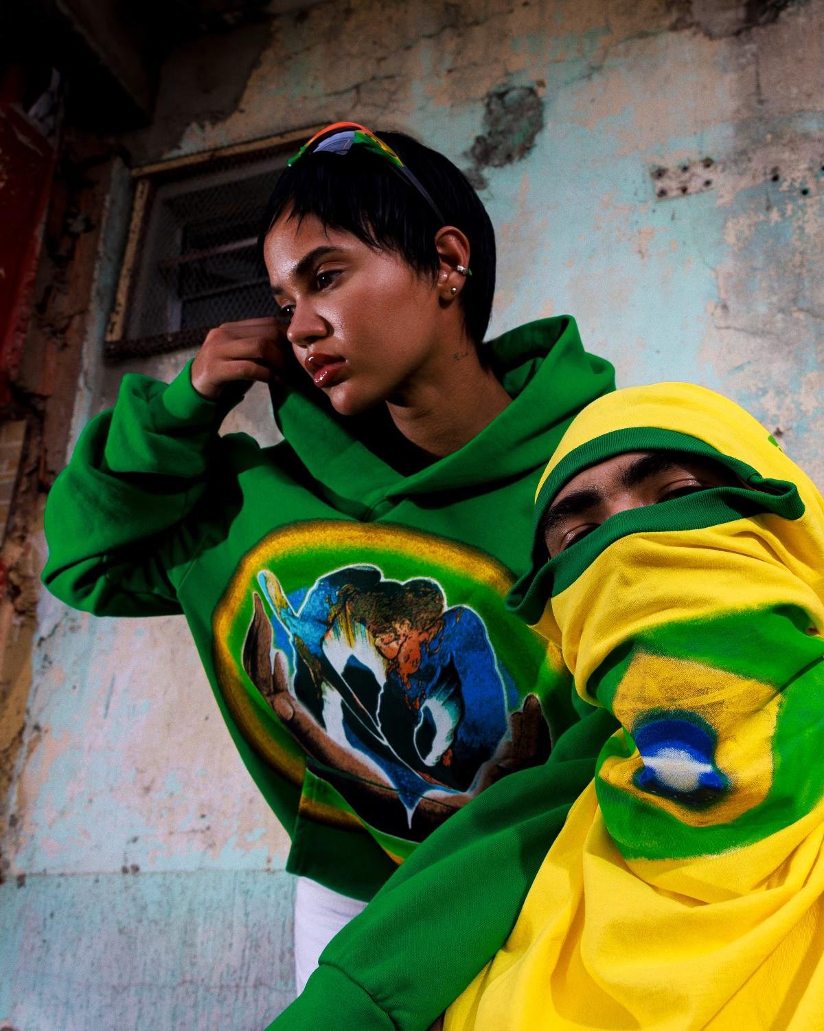 Moda Streetwear Brasileira: Imagem mostra duas pessoas vestindo moletons verdes com estampas artísticas e coloridas. Uma delas cobre o rosto com tecido amarelo e verde, combinando com óculos de sol, evidenciando uma tendência urbana e ousada.