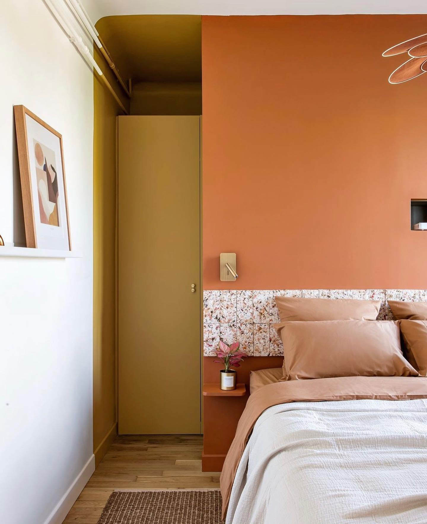 Quarto decorado em tons terrosos e detalhes florais, com paredes em terracota e mostarda. Móveis minimalistas e quadros de arte abstrata complementam o ambiente. Tendências de design escandinavo e uso de cores quentes para criar um espaço acolhedor.