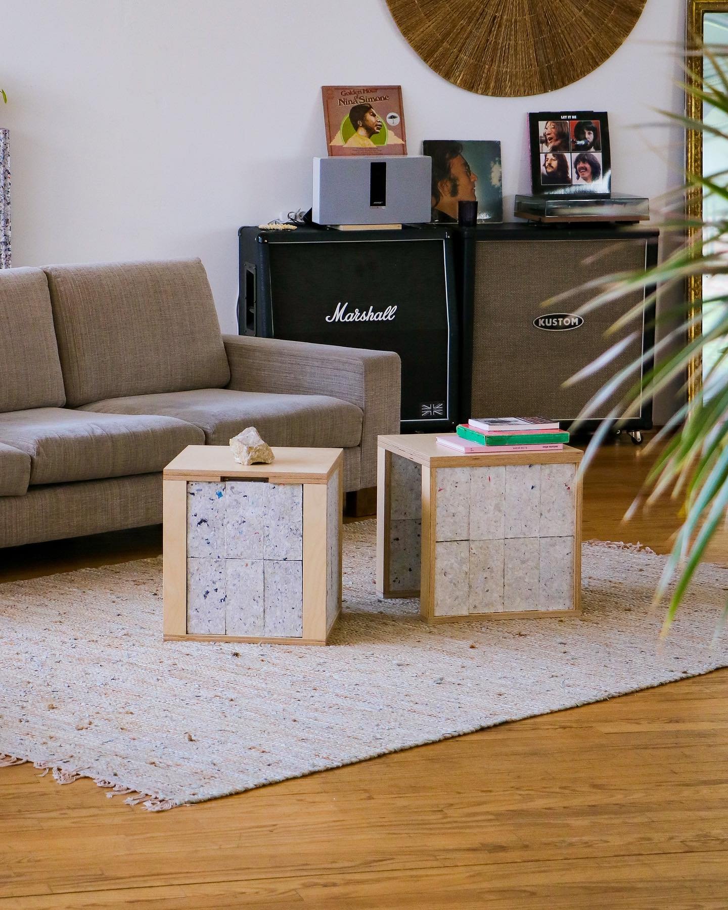 Sala de estar com decoração moderna e aconchegante. Sofá cinza claro, amplificadores Marshall e Kustom, discos de vinil, mesa de centro com cubos minimalistas de madeira, tapete de tecido natural e detalhes rústicos e musicais.