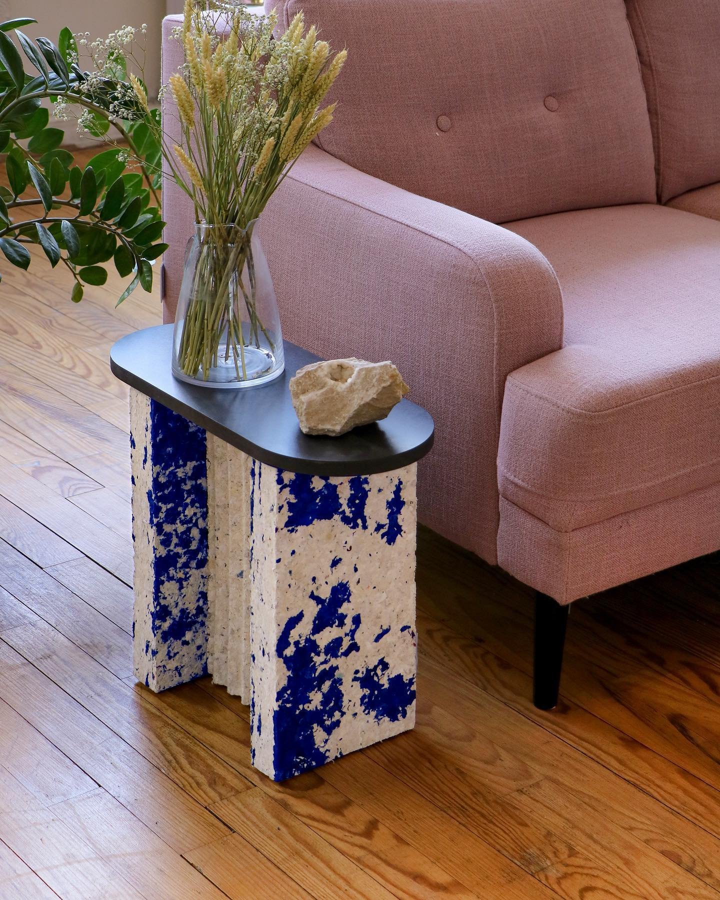 Sala de estar com estilo moderno. O sofá rosa segue a tendência de cores suaves, combinado com uma mesa lateral artística com base em textura pintada de azul e branco. O vaso com flores secas e a pedra decorativa adicionam um toque natural ao ambiente.