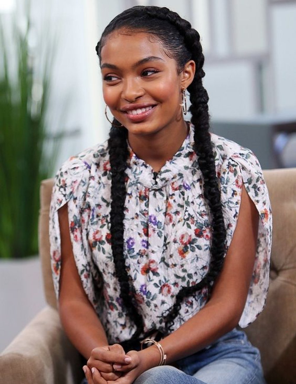 Mulher sorridente com estilo casual, usando uma blusa floral de manga curta e jeans. A trança boxeadora adiciona um toque moderno e descontraído. A moda floral e as tranças continuam sendo tendências populares, destacando a elegância simples.