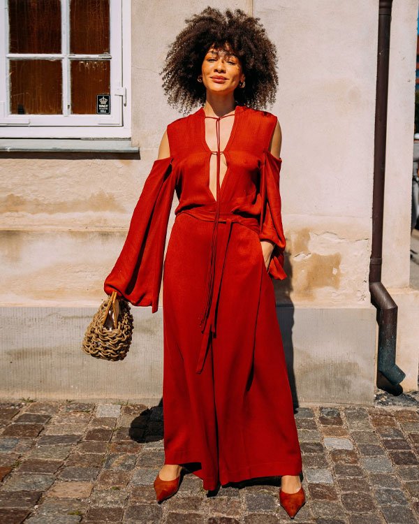 Mulher com vestido longo vermelho e elegante, com ombros à mostra e mangas largas. Ela segura uma bolsa de palha artesanal e usa sapatos de salto combinando com a cor do vestido.