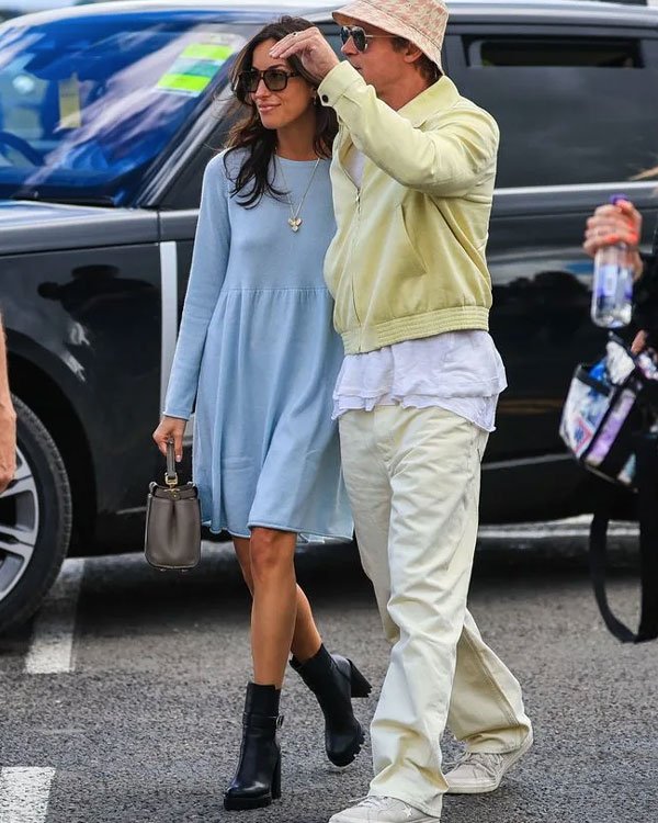 Casal usando tendências modernas: ela veste um vestido azul claro solto, botas pretas de cano curto e uma bolsa pequena. Ele combina jaqueta bege e chapéu em estilo casual, calça clara e tênis brancos.