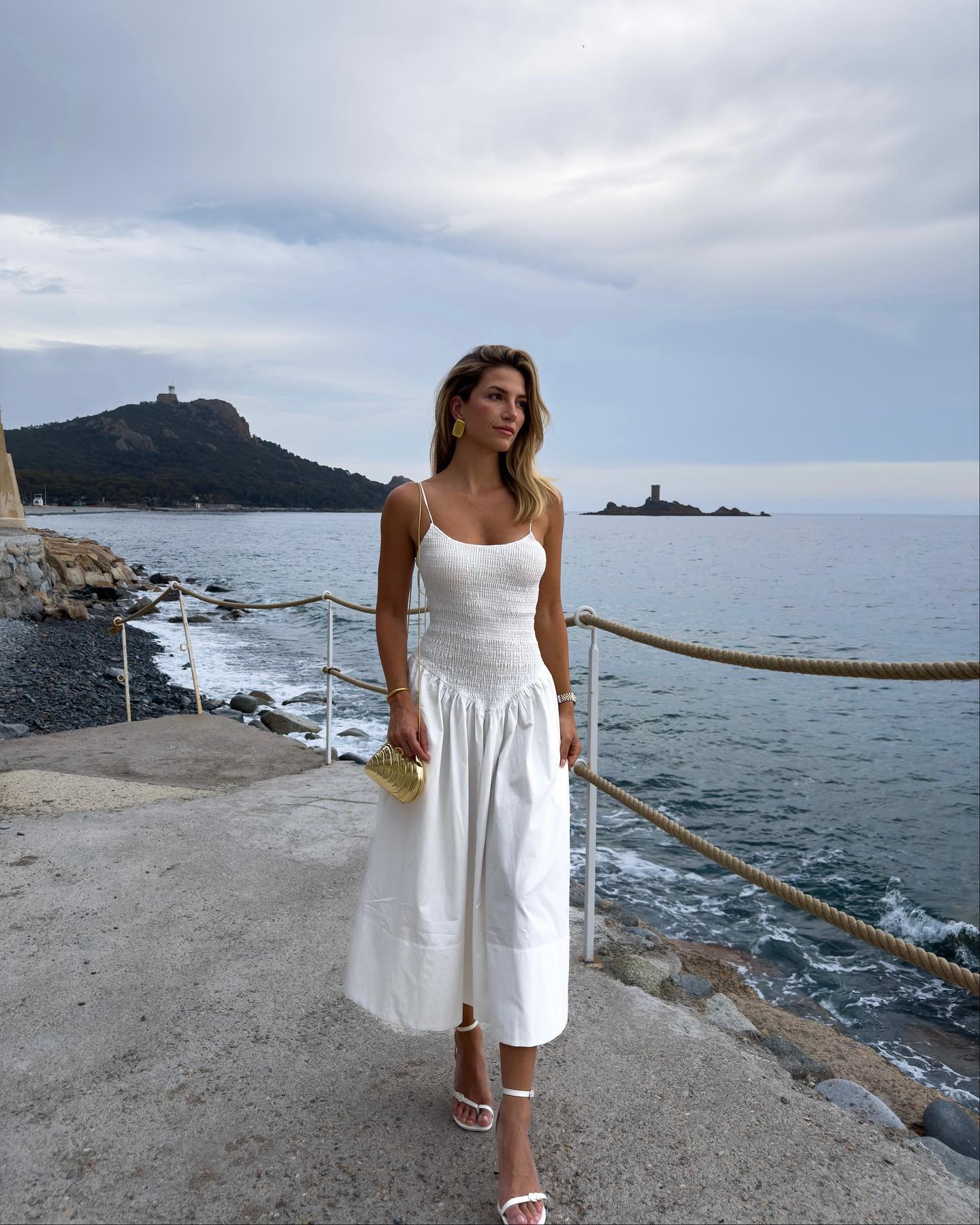 Mulher à beira-mar usando vestido branco midi com alcinhas finas e textura franzida. Complementa o look com uma sandália branca de salto baixo e uma bolsa dourada. Estilo elegante e minimalista, perfeito para um passeio ao ar livre.