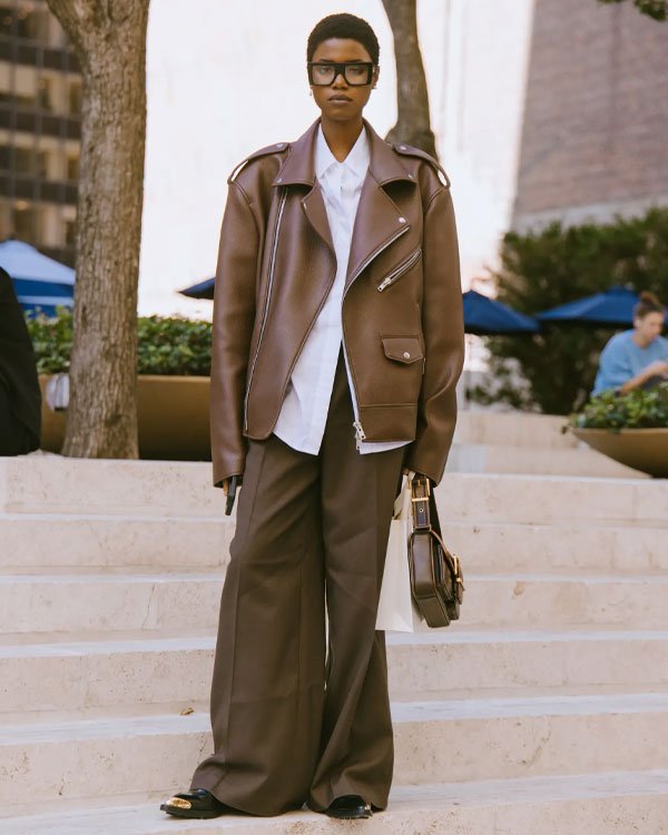 Pessoa vestindo jaqueta oversized marrom, camisa social branca e calça larga marrom. O look é arrematado por óculos grandes de armação preta e bolsa pequena de couro.