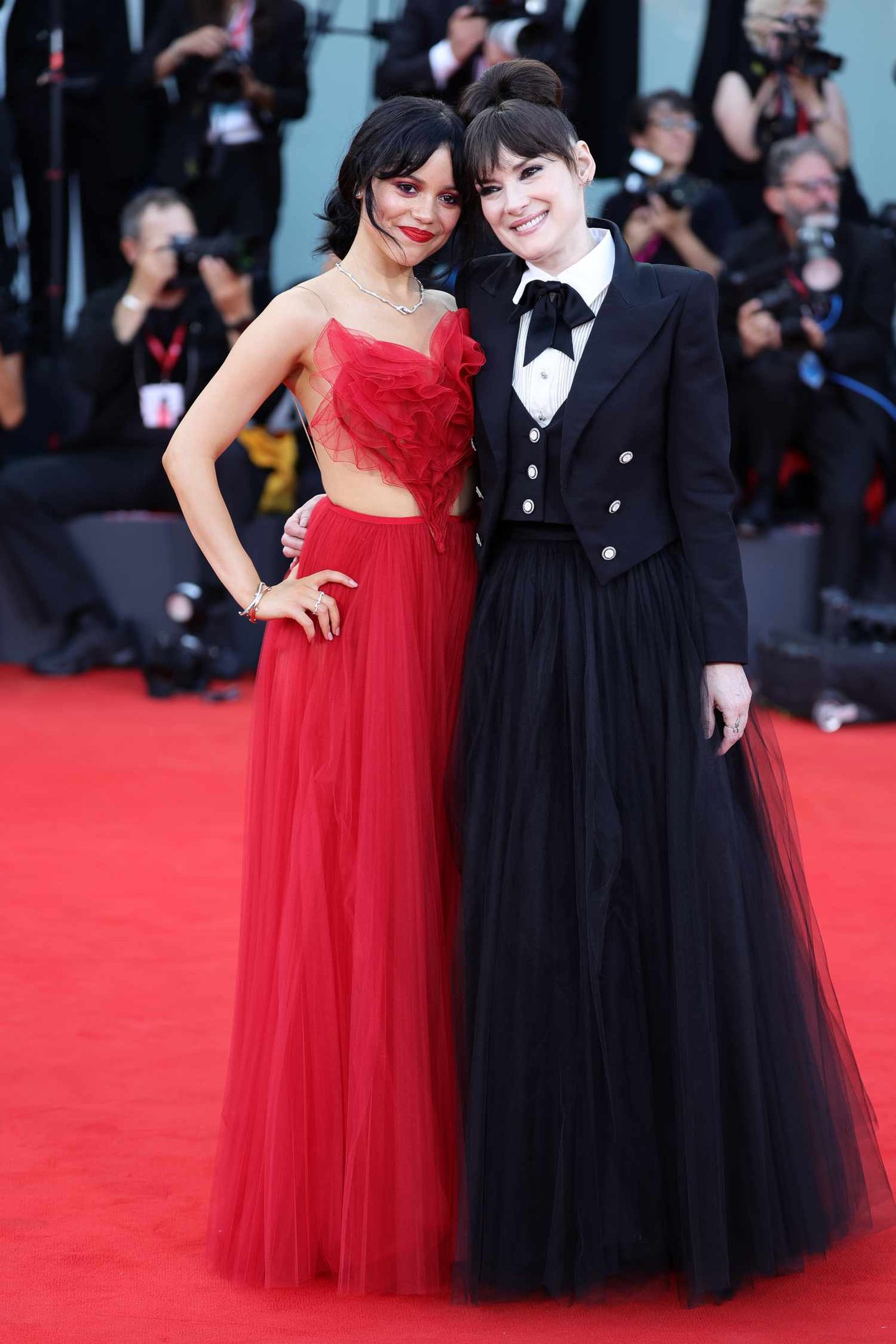 Duas mulheres posando no tapete vermelho, uma com vestido de tule vermelho vibrante e a outra com traje preto estiloso composto por blazer, camisa branca e saia longa de tule preto.