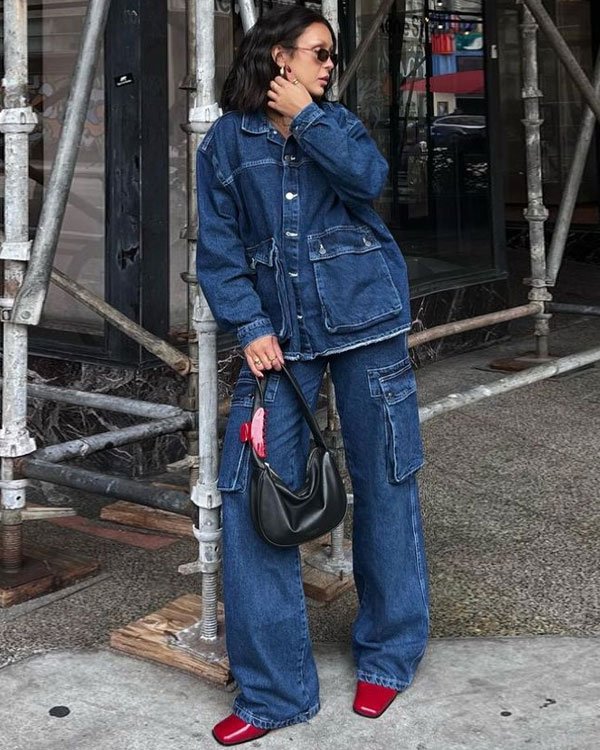 Pessoa vestida estilo urbano: conjunto oversized de jaqueta e calça jeans de lavagem escura com bolsos grandes, sapatos vermelhos, óculos de sol e bolsa preta destacam as tendências de moda casual e utilitária.