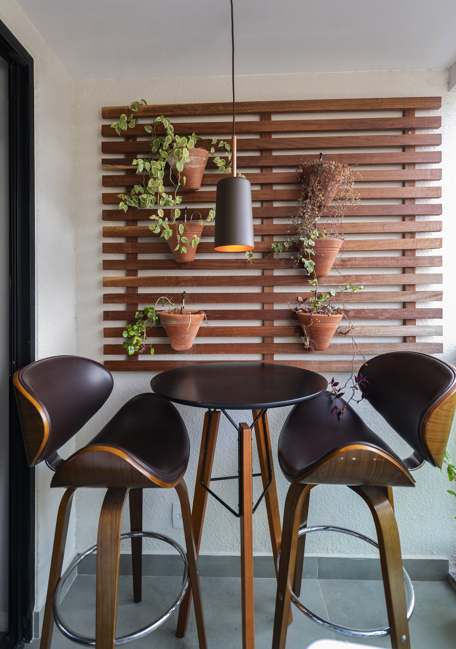 Mesa com três cadeiras em varanda de apartamento com plantas em vasos suspensos na parede de madeira.