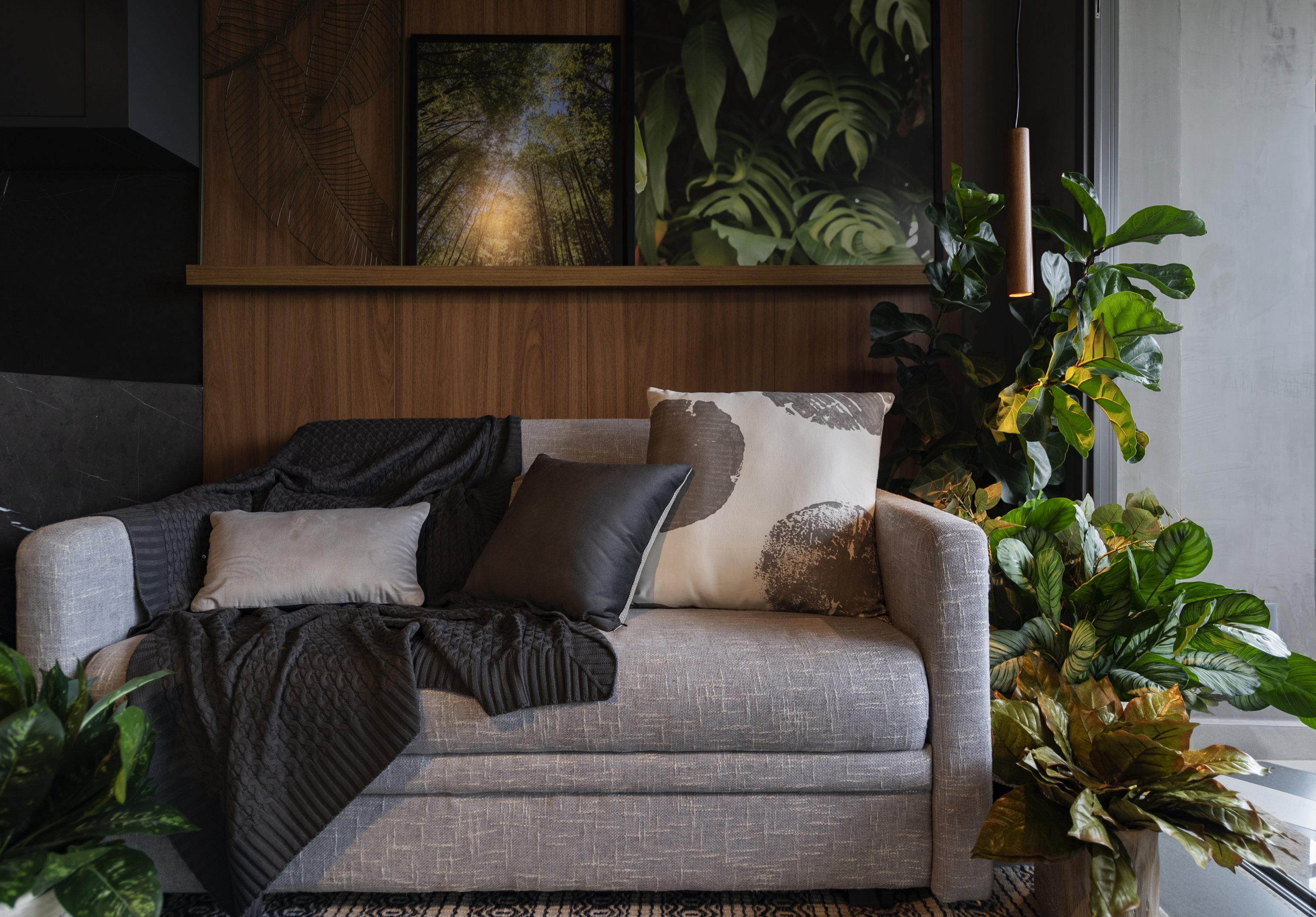 Sala de estar em um apartamento com sofá cinza, almofadas decorativas e várias plantas ao redor.