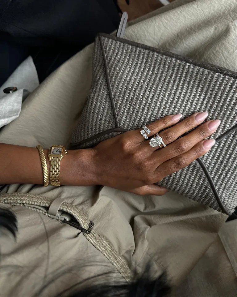 Mão adornada com anéis de diamante, relógio dourado estilo retrô e bolsa texturizada.