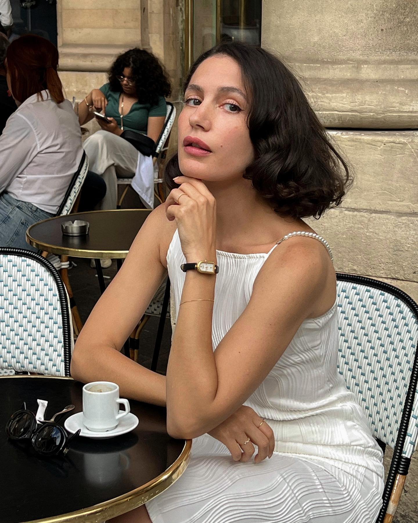 Mulher com vestido branco em um café ao ar livre, usando um relógio de pulso clássico. óculos de sol na mesa.