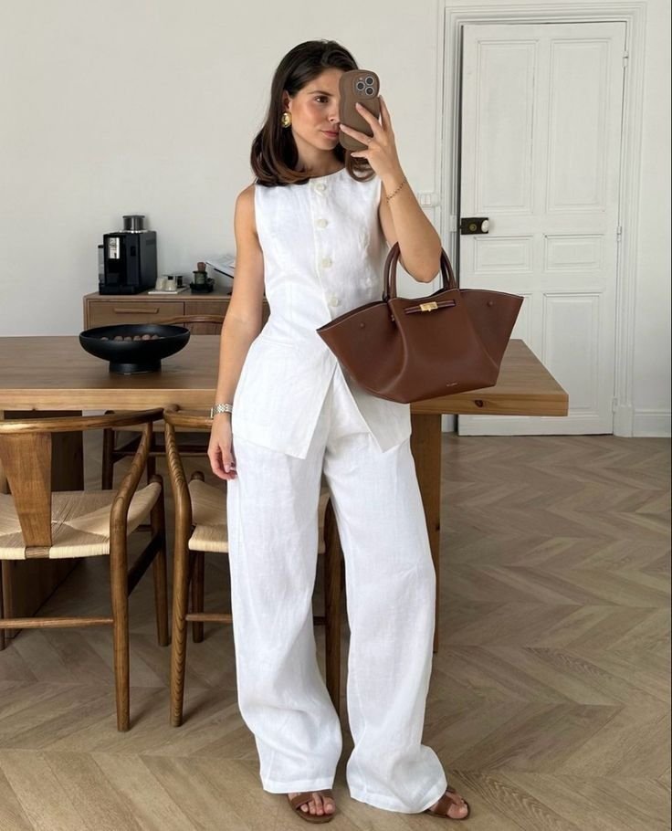 Mulher com conjunto de linho branco, composto por blusa sem mangas e calça ampla. Acessórios incluem brincos dourados, smartwatch e sandálias marrons. Completa o look casual e elegante com bolsa grande marrom.