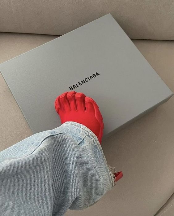 Calça jeans com caimento largo e sapato vermelho de dedos separados, estilo inovador da Balenciaga, destacando tendências de moda ousadas e modernas. Combinação audaciosa de tonalidades e design.