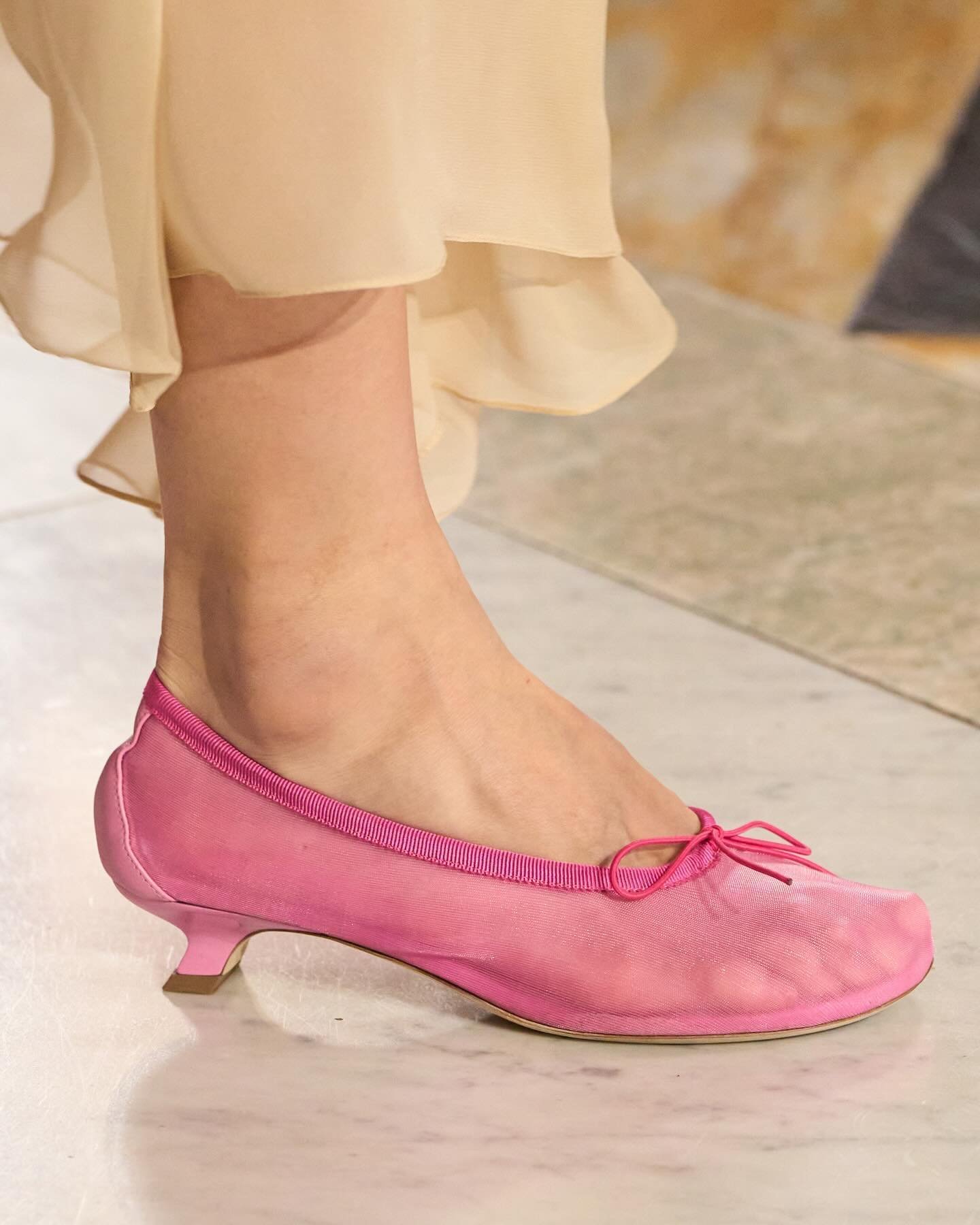 Sapato rosa com salto baixo estilo kitten heel e laço delicado, combinado com vestido de tecido leve e fluido. Representa tendência moderna de conforto e elegância em looks femininos, unindo tradição e sutilidade.