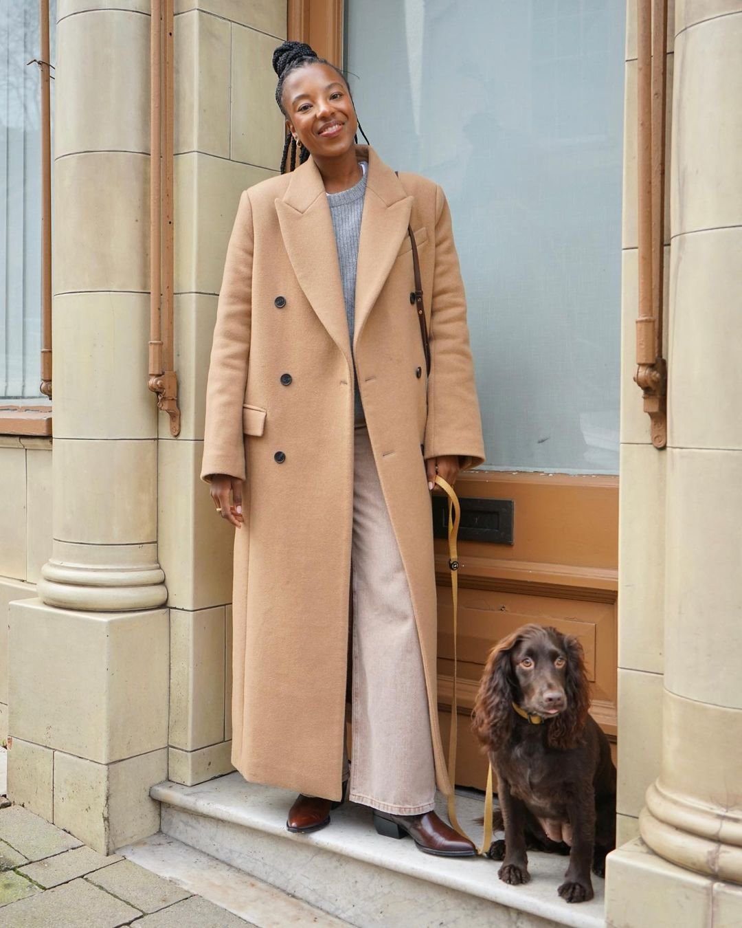 Mulher sorridente usando sobretudo bege, calça clara e botas marrons, segurando a guia de um cachorro marrom em uma entrada de prédio.