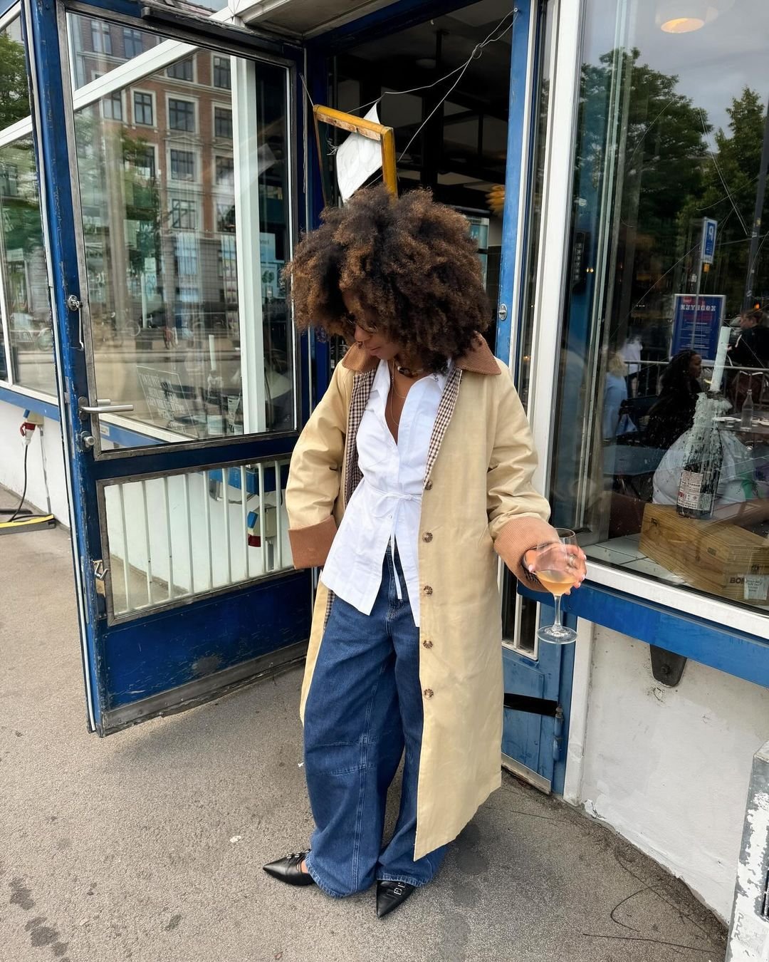 Mulher com sobretudo bege, camisa branca e calça jeans larga, segurando uma taça de bebida, em frente a um restaurante com estrutura azul.