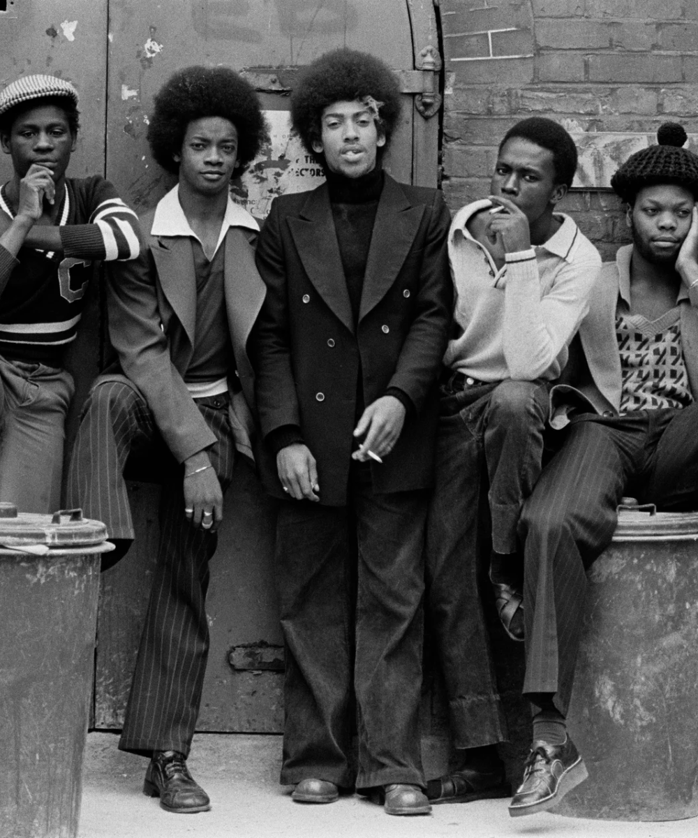 Jovens exibem estilo vintage dos anos 1970, com cabelos afro, paletós de lapela larga, suéteres, calças com listras e boinas. O visual mistura elegância e casualidade, refletindo tendências urbanas da época.