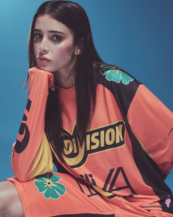 Livia e (di)vision. Modelo veste camisa oversized laranja, com estampas gráficas e coloridas, tendência streetwear. O visual mistura elementos esportivos e contemporâneos, destacando-se pela ousadia e estilo urbano.