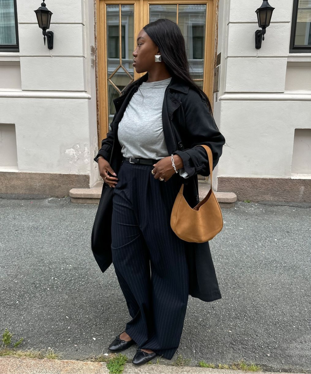 Mulher usando casaco preto e blusa cinza, segurando bolsa fashionista caramelo, posando em frente a uma porta de madeira.