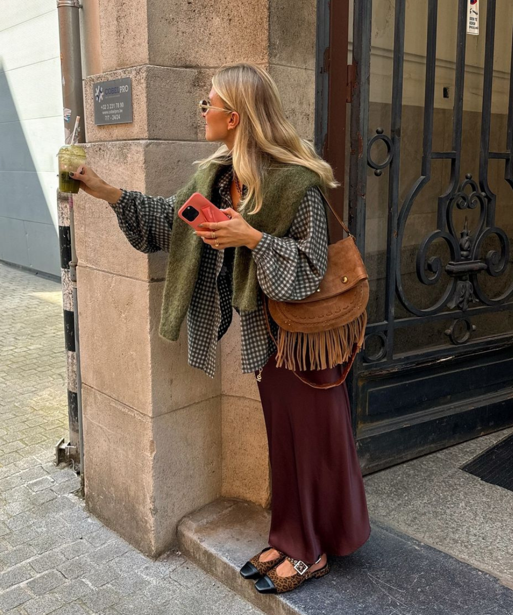 Mulher com casaco verde e saia longa, segurando bebida e celular, usa bolsa com franjas.