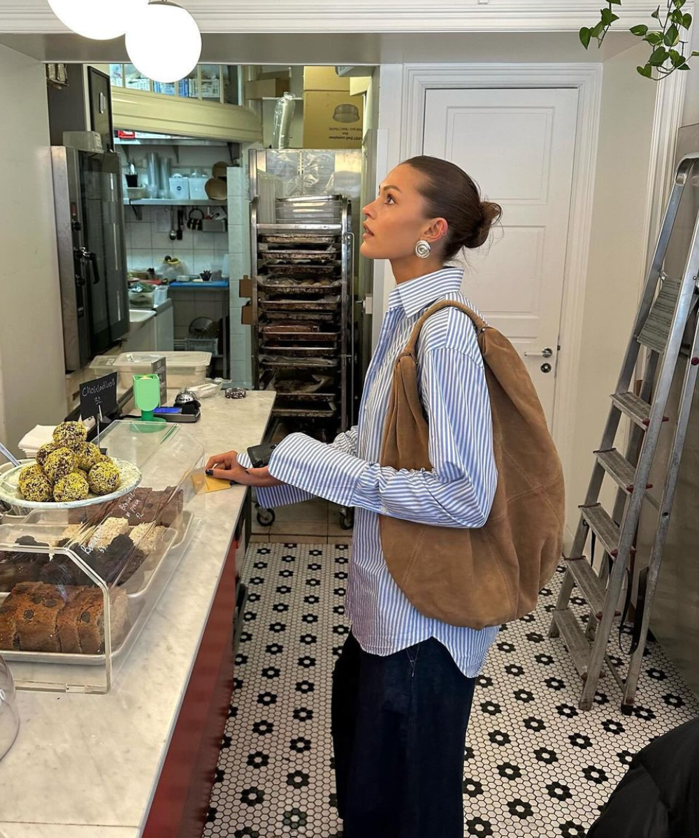 Mulher em padaria usando camisa listrada e bolsa fashionista de camurça marrom, observando itens no balcão.