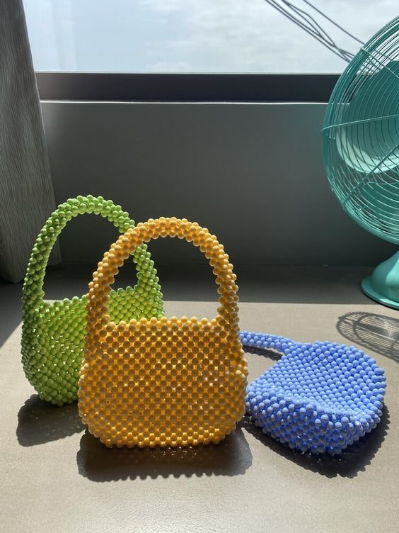 Bolsas de contas coloridas em verde, amarelo e azul, refletindo tendências contemporâneas de moda artesanal e acessórios vibrantes. Estilo divertido e casual, ideal para looks despojados e modernos.