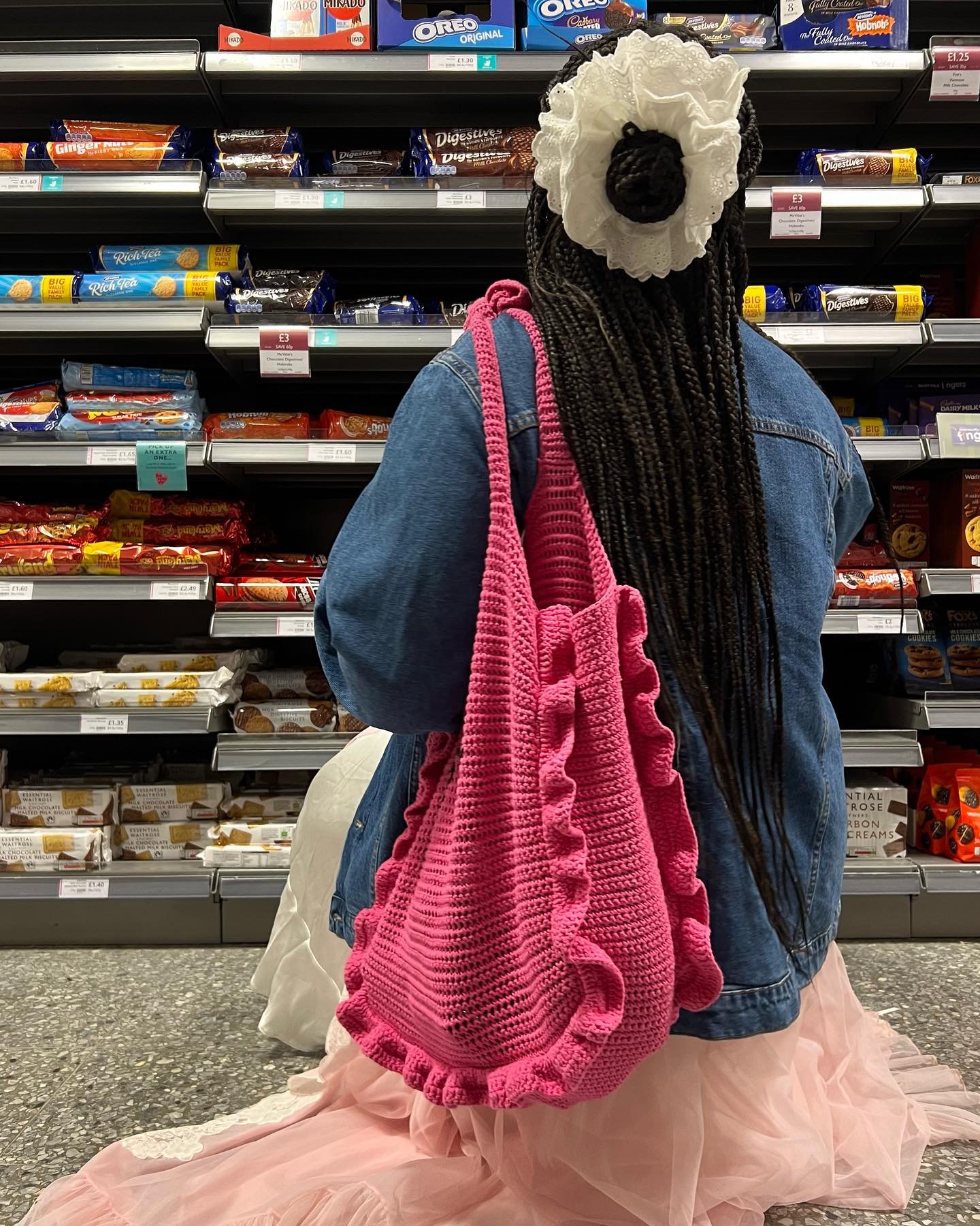 Pessoa usando jaqueta jeans, saia rosa fluida e bolsa de crochê rosa. Destaque para scrunchie de renda no cabelo. A combinação mistura estilos retrô e moderno, enfatizando conforto e criatividade em moda casual.