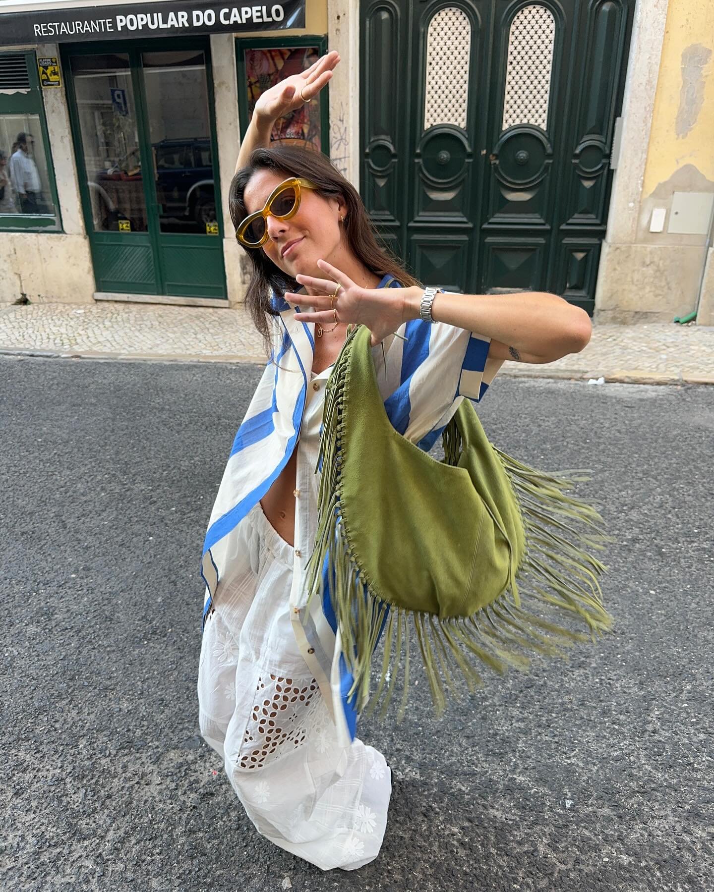 Despojado e moderno, com camisa listrada, saia branca rendada e óculos de sol estilo retrô. A bolsa verde com franjas adiciona um toque boho ao look urbano.