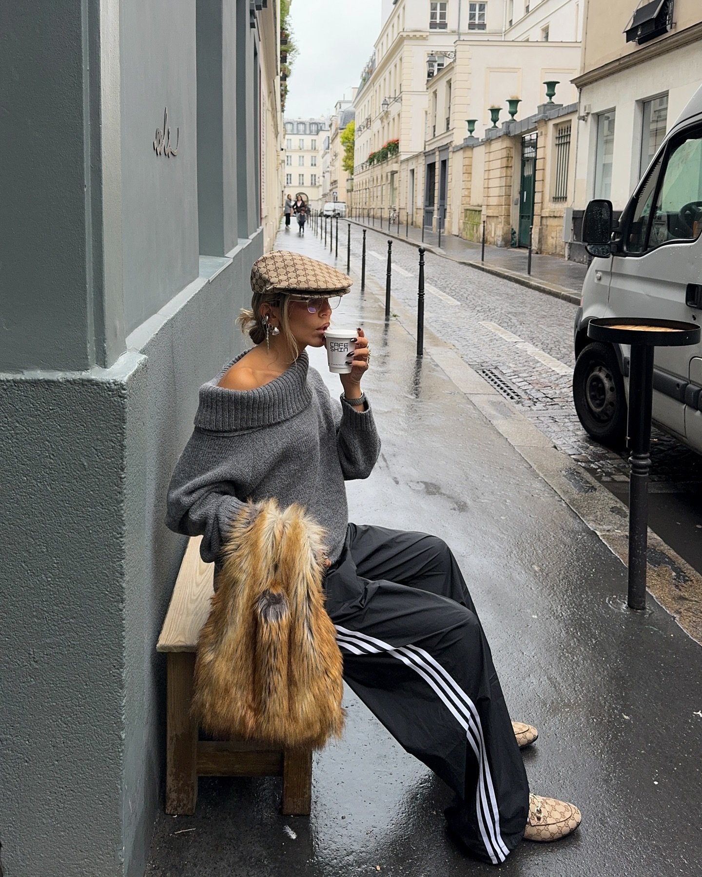 Pessoa sentada em Paris, usando suéter cinza oversized e boina xadrez. Calça esportiva com listras, bolsa de pele sintética e mocassins completam o look casual e urbano. Tendência de misturar peças esportivas com toque clássico.