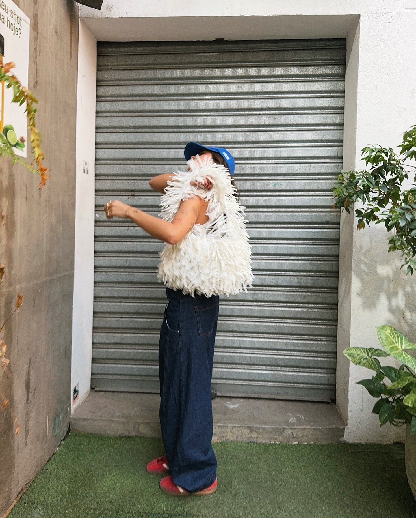 Pessoa usando colete felpudo branco, calça jeans larga e tênis vermelho. Completa o look com boné azul, em ambiente urbano. Combinação de texturas e tons vibrantes em estilo casual moderno.