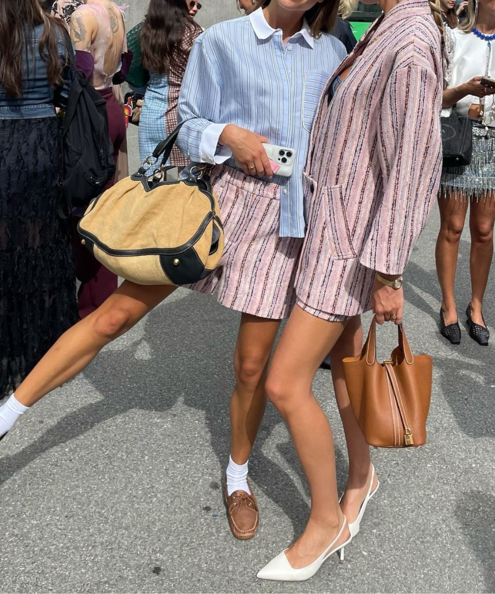 Duas mulheres com looks elegantes em tons pastel e listras. Uma usa camisa azul e bolsa bege; a outra, conjunto rosa e bolsa marrom. Sapatos variados, dos clássicos aos modernos, refletem tendências casuais e sofisticadas.