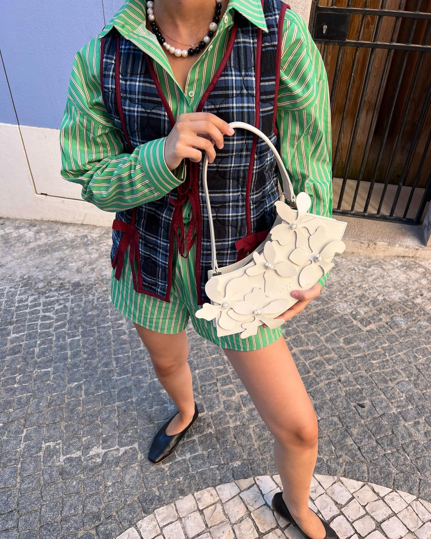 Look eclético com camisa listrada verde e colete xadrez azul. Shorts combinando. Bolsa branca com detalhes florais e sapatos pretos minimalistas. Colar de pérolas destaca o visual. Tendências de mix de estampas e acessórios ousados.