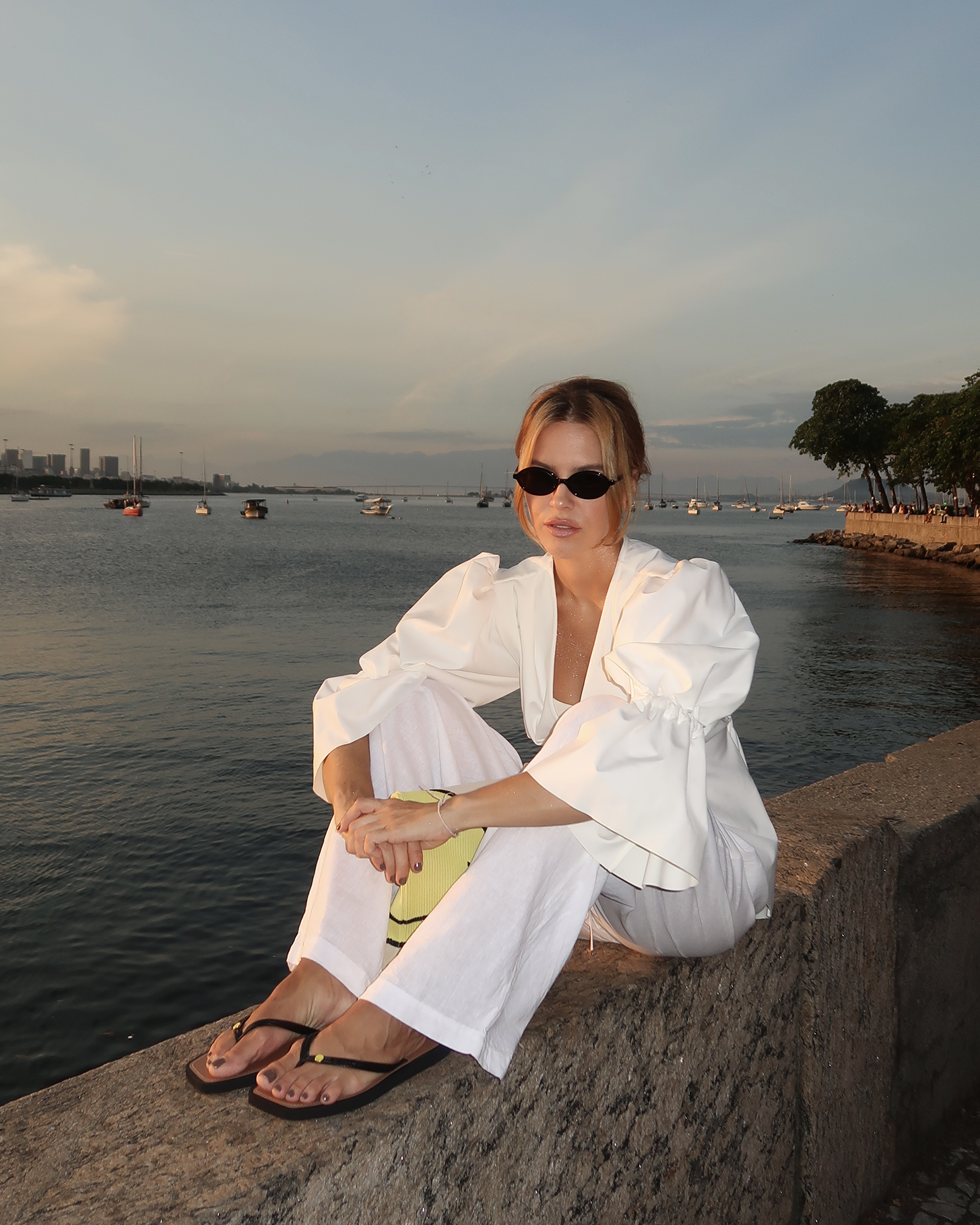 Pessoa com óculos escuros veste roupa branca e senta à beira-mar ao pôr do sol, segurando uma bolsa amarela com logo Smiley. Brizza e Smiley