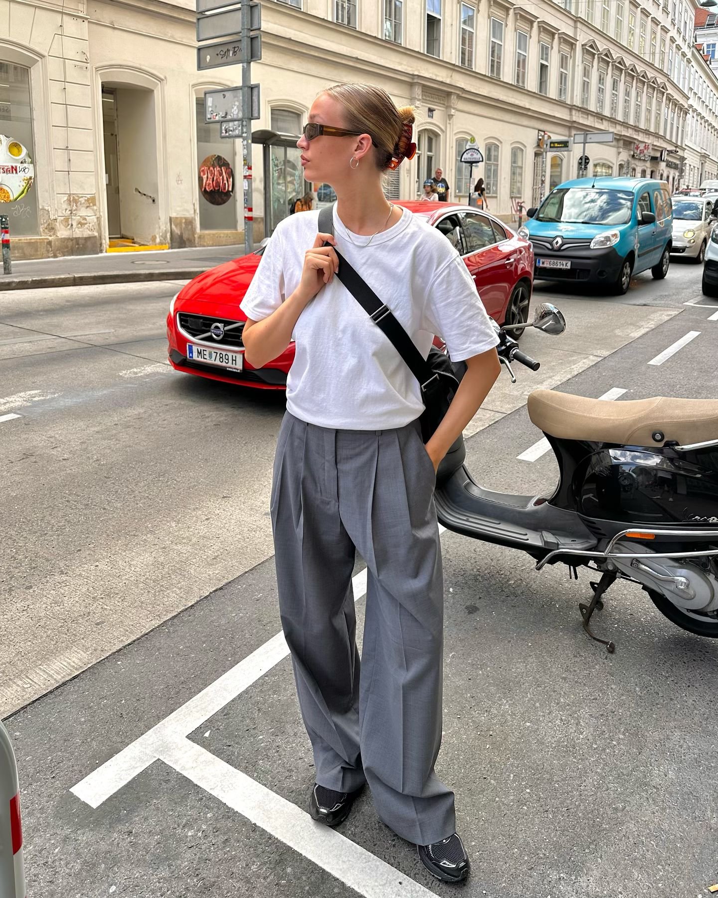 Mulher de camiseta branca e calça da moda cinza larga em rua movimentada, ao lado de scooter e carro vermelho. Usa óculos escuros.