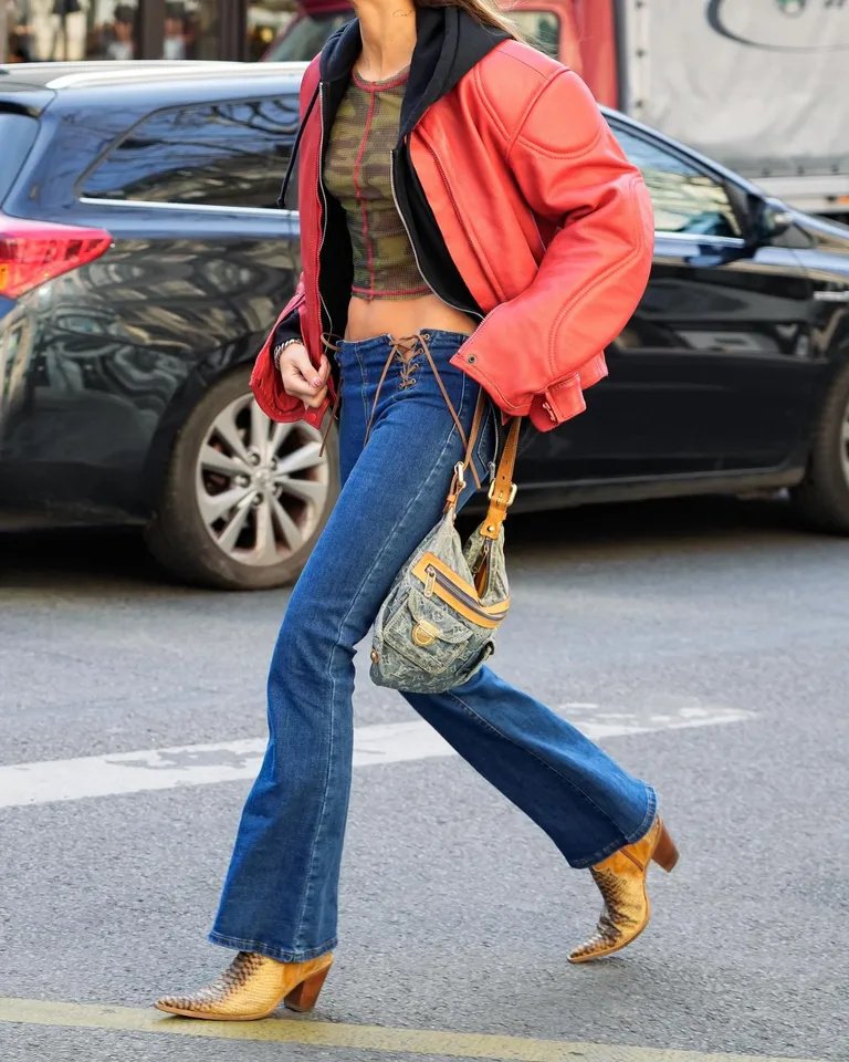 Pessoa usando jaqueta oversized vermelha, top camuflado, calça jeans de cintura baixa com corte flare e botas de couro com estampa, complementadas por uma bolsa estilo vintage. Look mistura tendências retrô e modernas.