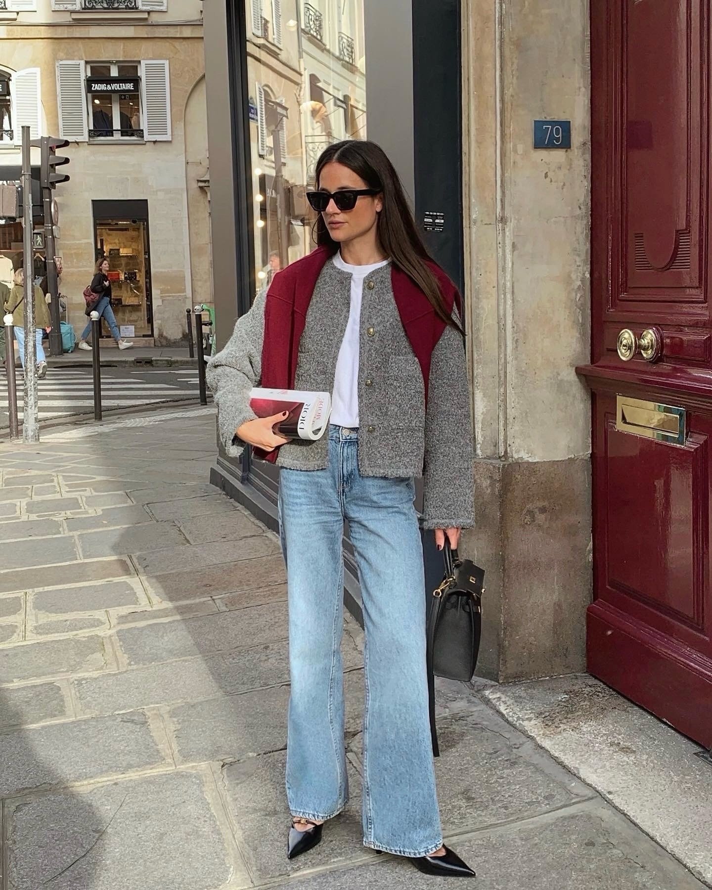 Mulher com look casual-chique em calça da moda jeans, camiseta branca e casaco cinza. Suéter vermelho nos ombros. Acessórios incluem bolsa preta, livro e óculos escuros, combinando elegância e descontração urbana em tendência moderna.