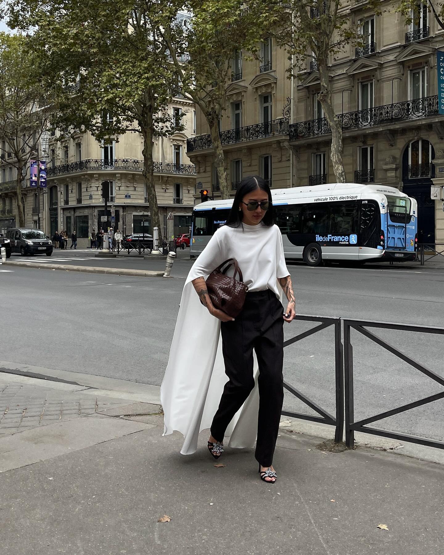 Pessoa usando capa branca longa, blusa de gola alta, calça preta e sandálias de salto baixo com pedrarias, em cenário urbano. Look sofisticado e minimalista em tendência, com bolsa marrom grande e óculos escuros.