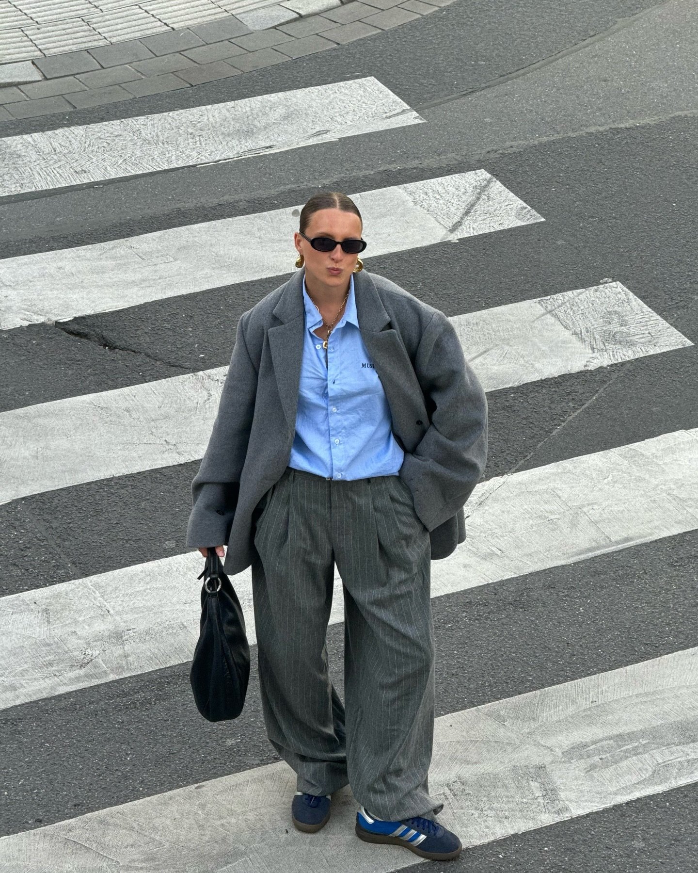 Pessoa atravessa a rua com look elegante e despojado: blazer oversized cinza, camisa azul, calça ampla e tênis. Óculos escuros e bolsa preta completam o visual urbano, destacando tendências de moda casual e minimalista.