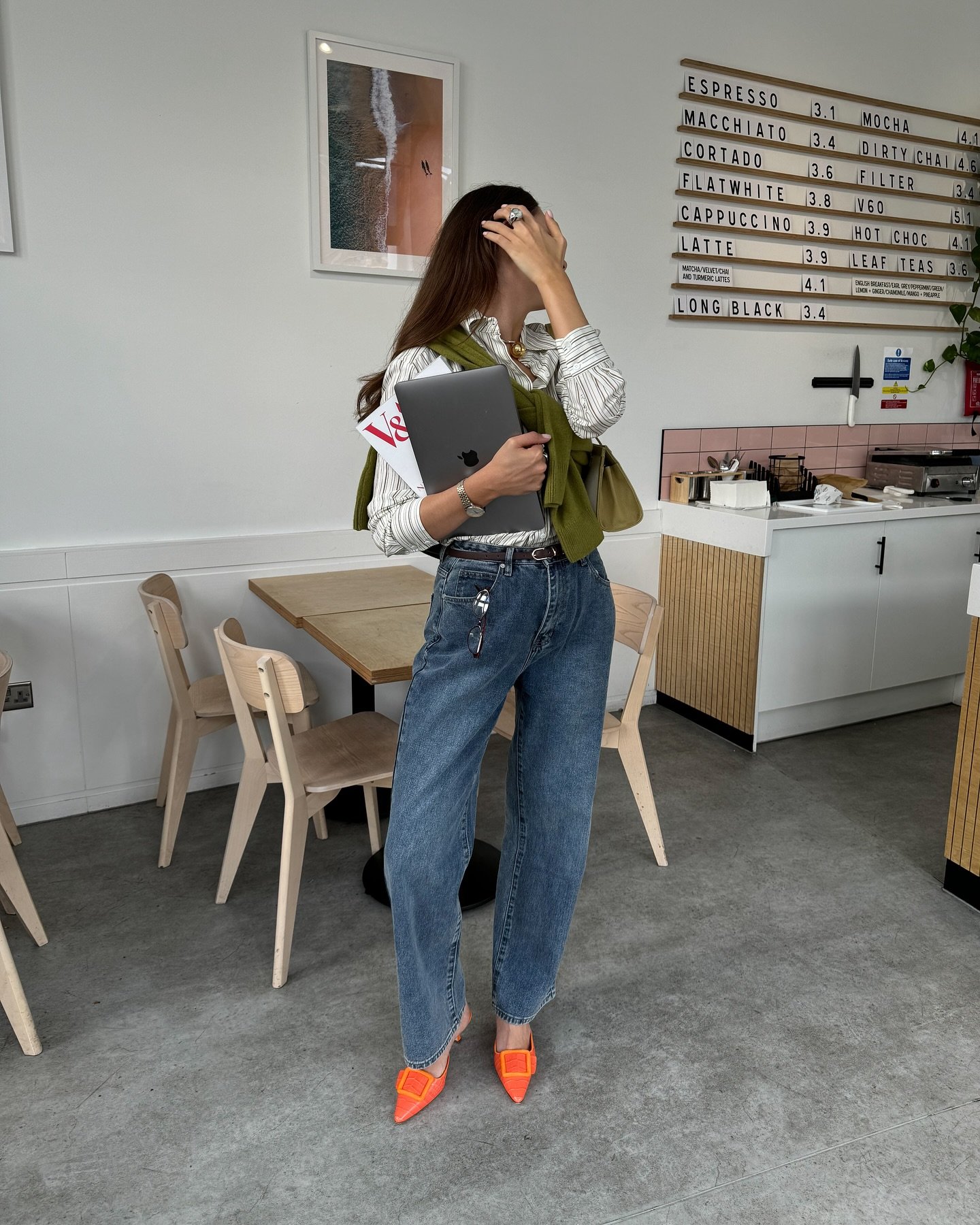 Mulher em café, vestindo camisa listrada, suéter verde sobre os ombros, calça jeans ampla e sapatos laranja vibrante. Look casual e moderno, destacando tendências de conforto e estilo urbano. Acessórios discretos completam o visual.