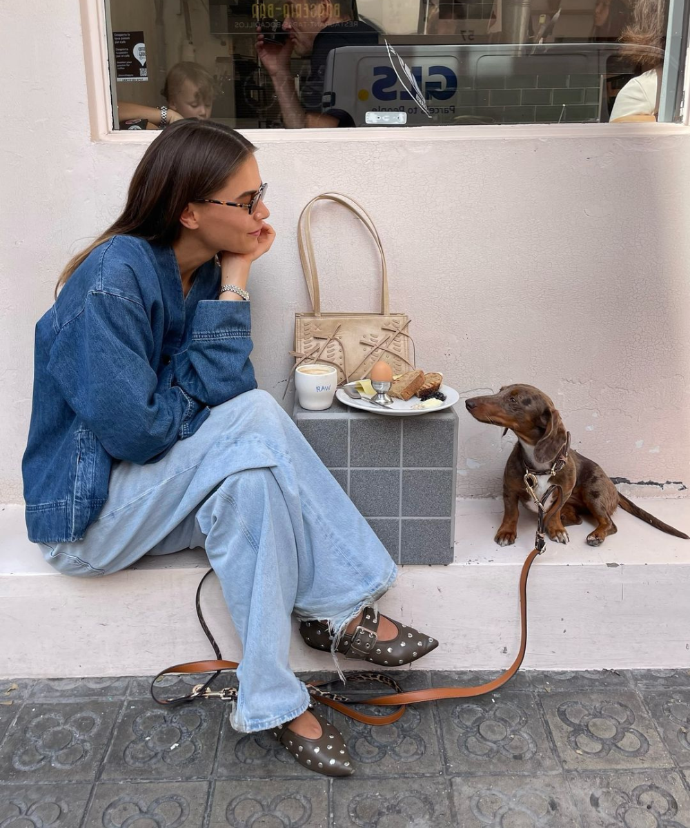 Mulher sentada casualmente com jaqueta oversized e jeans claros, destacando tendências de moda confortáveis. Sapatos escuros com detalhes de metal e bolsa bege completam o visual despojado, enquanto um cachorro pequeno está ao lado.