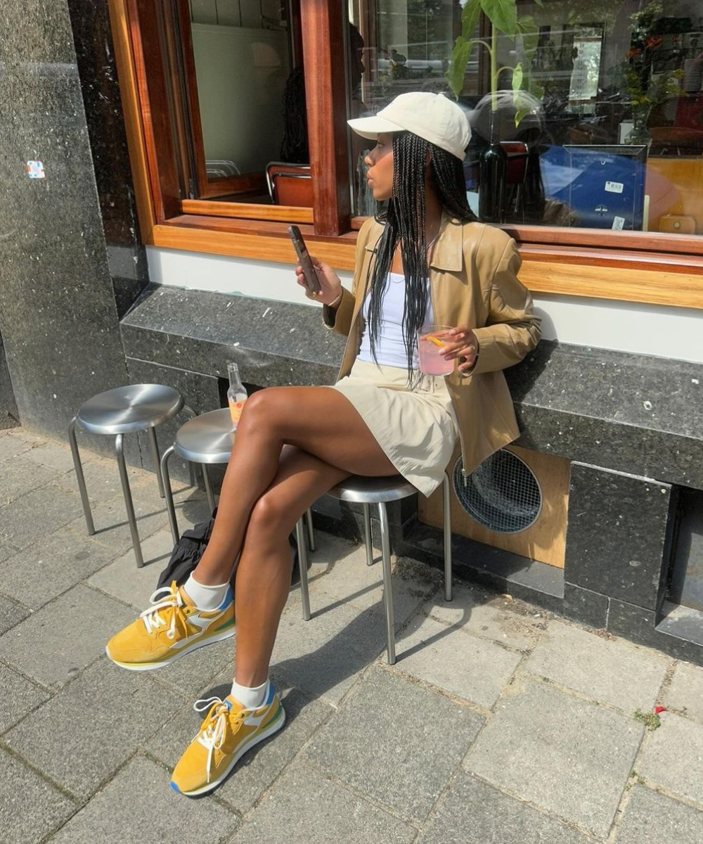 Mulher estilosa com look casual chic: blazer bege, top branco, saia curta e tênis amarelos vibrantes. Boné branco complementa o visual, trazendo descontração e sofisticação. Tendência urbana versátil e moderna.