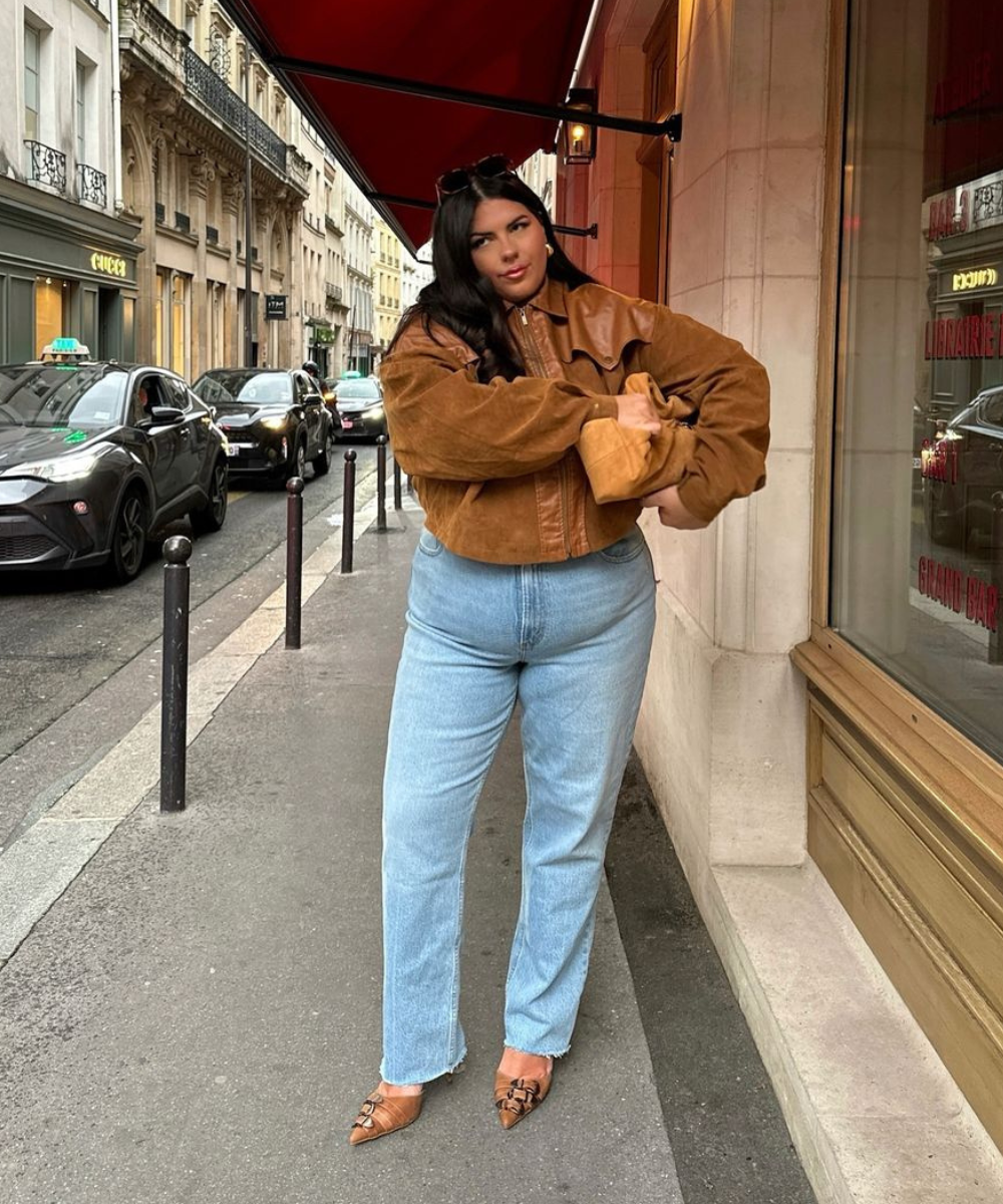 Mulher com jaqueta marrom oversized e calça jeans reta azul claro. Bolso retrô e sapatilhas estilo vintage. Look casual-chique urbano, destaque para os tons terrosos contrastando com o denim. Tendência aconchegante e elegante nas ruas da cidade.