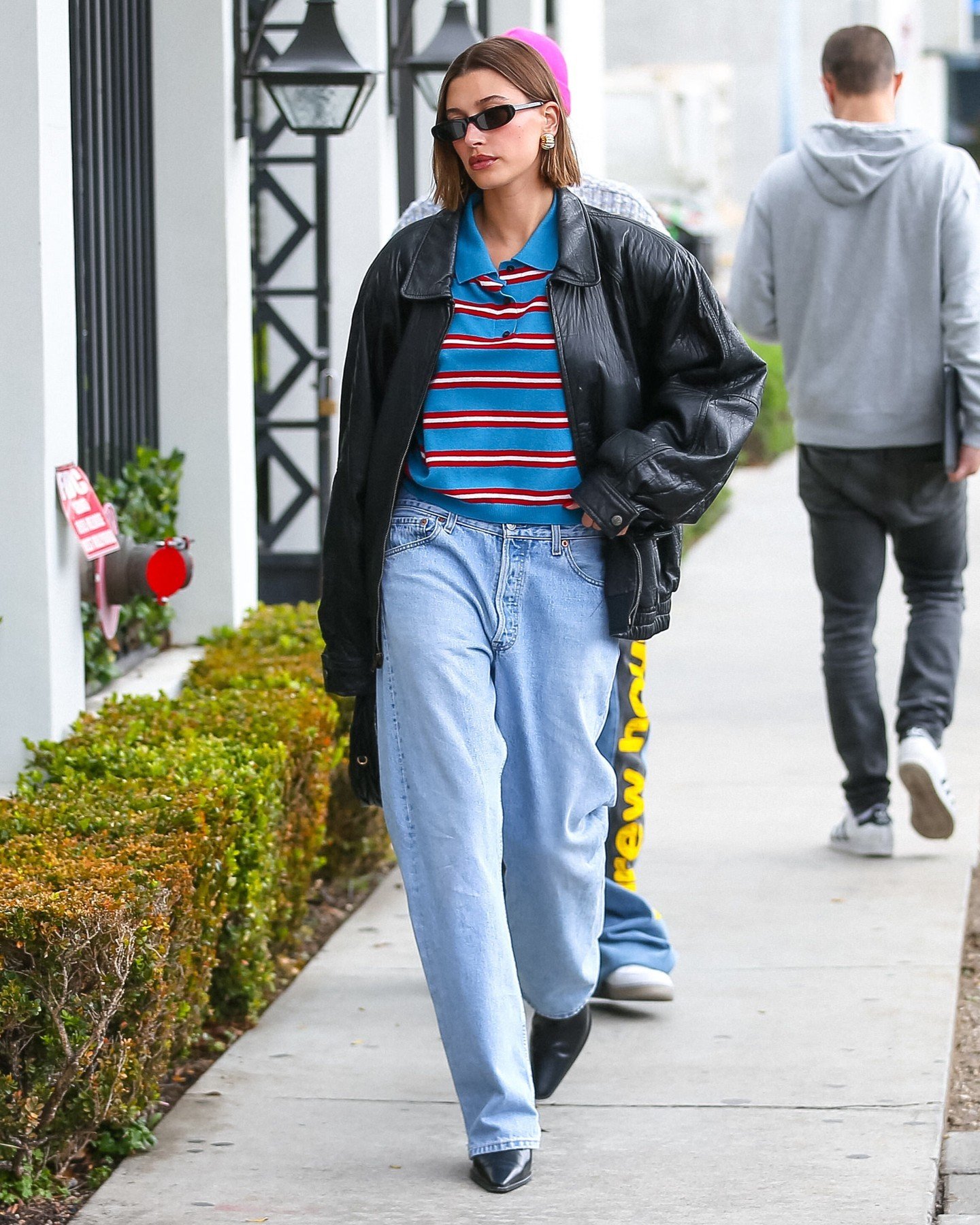 Mulher usando camisa polo listrada, jaqueta de couro preta e jeans largos, caminhando pela calçada.