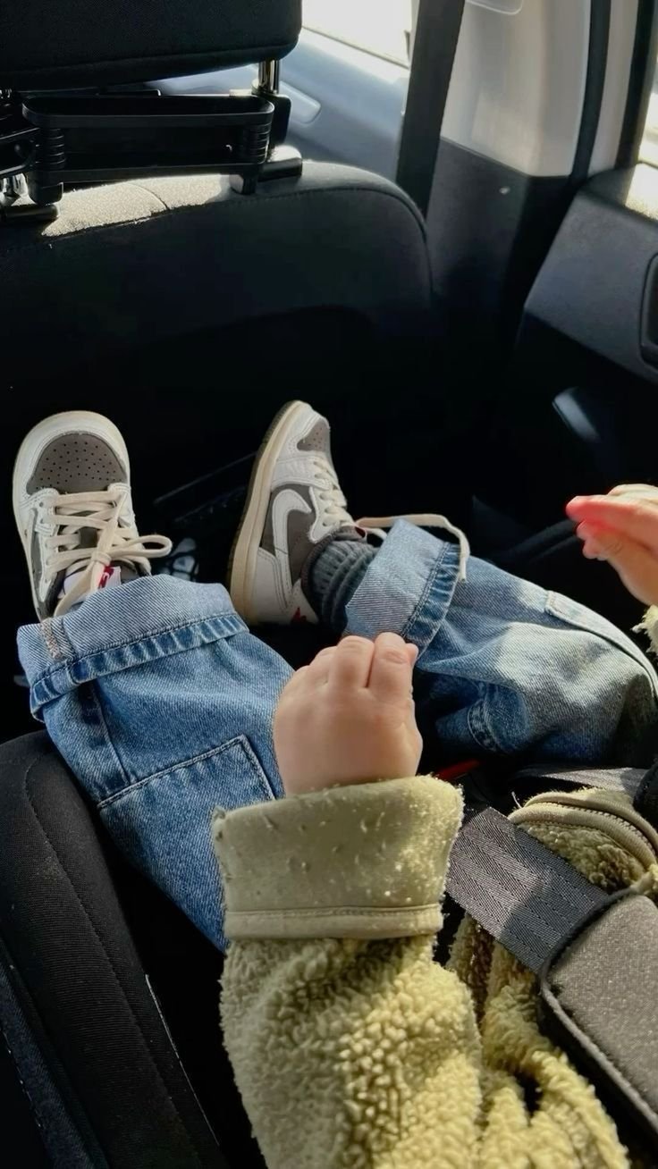 Criança no carro usando tênis esportivos brancos com detalhes em cinza, calça jeans larga dobrada na barra e casaco de lã bege. Look casual e confortável, perfeito para estilos infantis modernos. Bebê Conforto