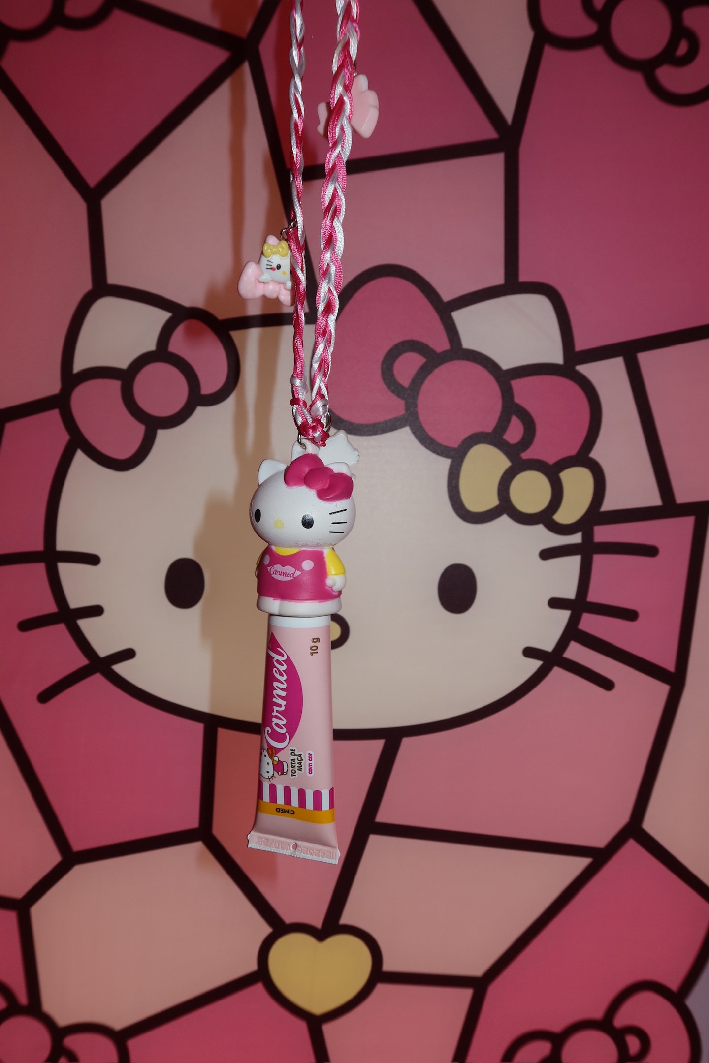 Carmed Hello Kitty pendurado em cordão trançado rosa, fundo com estampa da personagem.