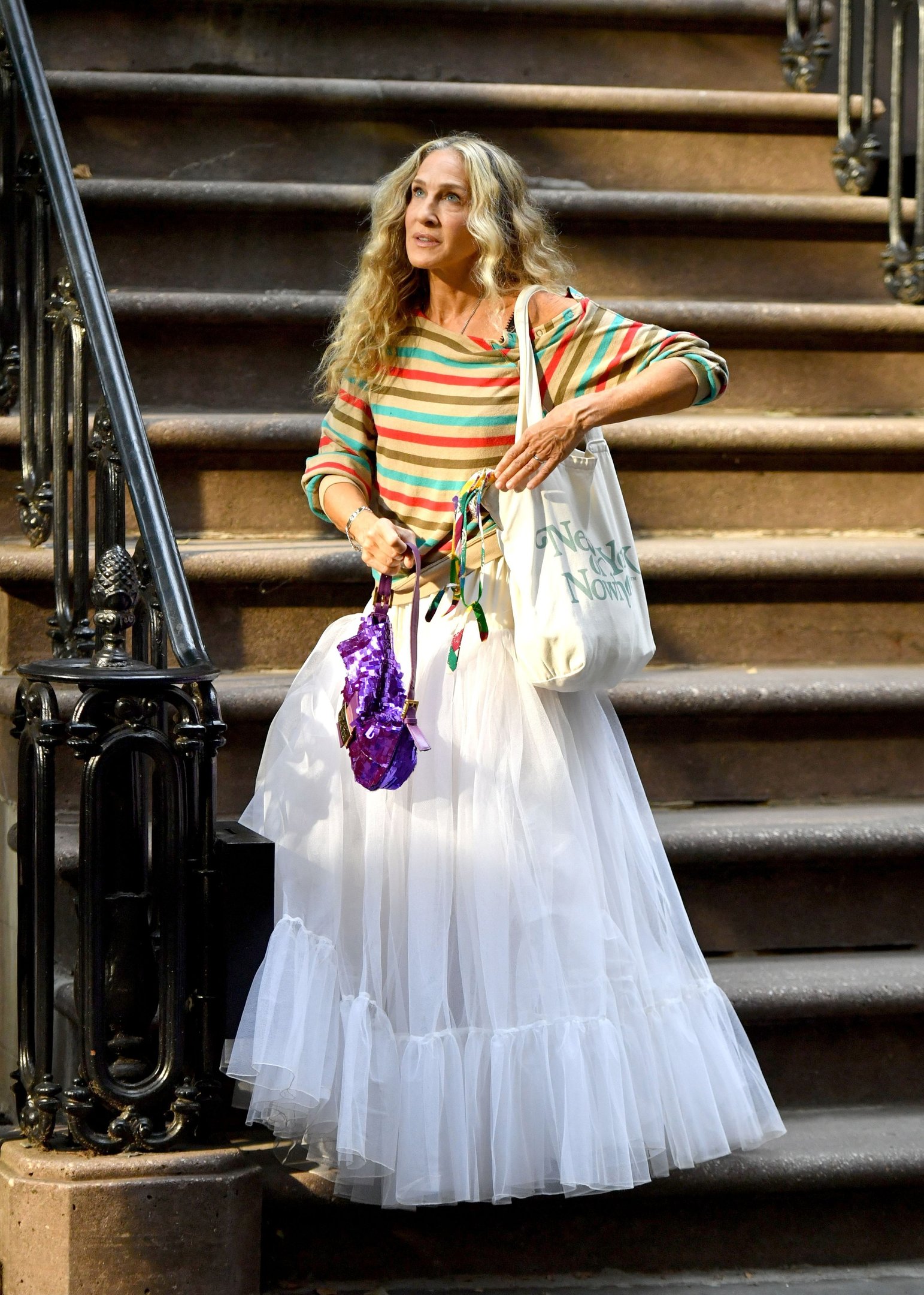 Mulher usando blusa listrada colorida e saia volumosa branca. O look combina casualidade e ousadia com bolsa de tecido e acessório roxo brilhante. Estilo eclético e retrô, sempre na moda.