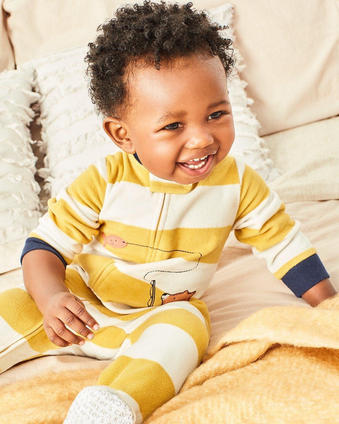 Bebê sorrindo sentado em um sofá, vestindo roupa de bebê listrada amarela e branca.