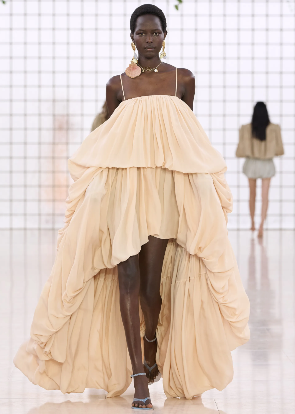 Modelo desfila usando um vestido de camadas volumosas em tom nude, com alças finas, destacando-se pela leveza e elegância fluida. Acessórios grandes e chamativos complementam o look, enquanto sandálias minimalistas acrescentam um toque de modernidade. Tendência: elegância etérea e uso de volumes.